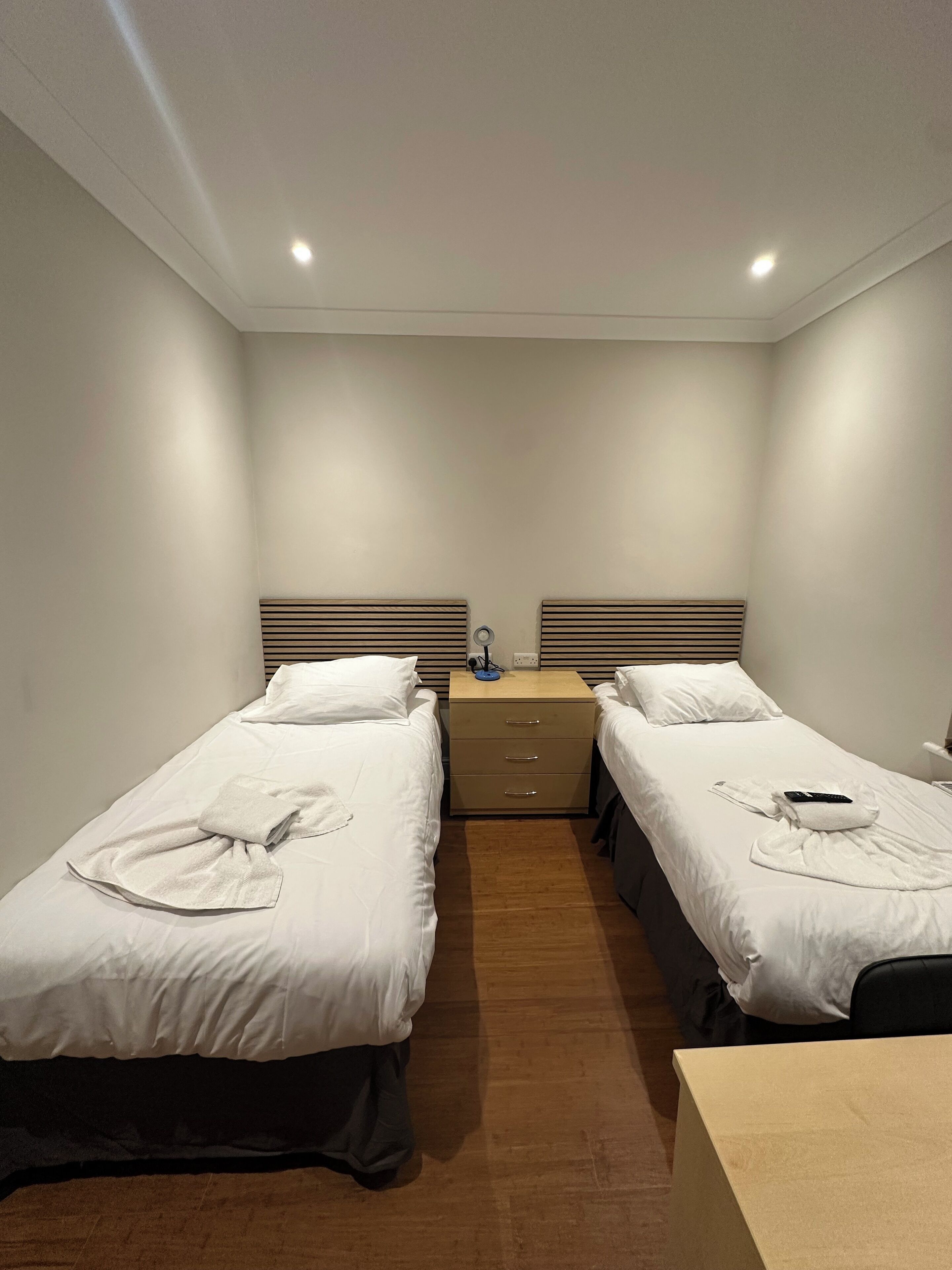 A hotel room with two single beds, a wooden nightstand with drawers, a desk, and a small table.