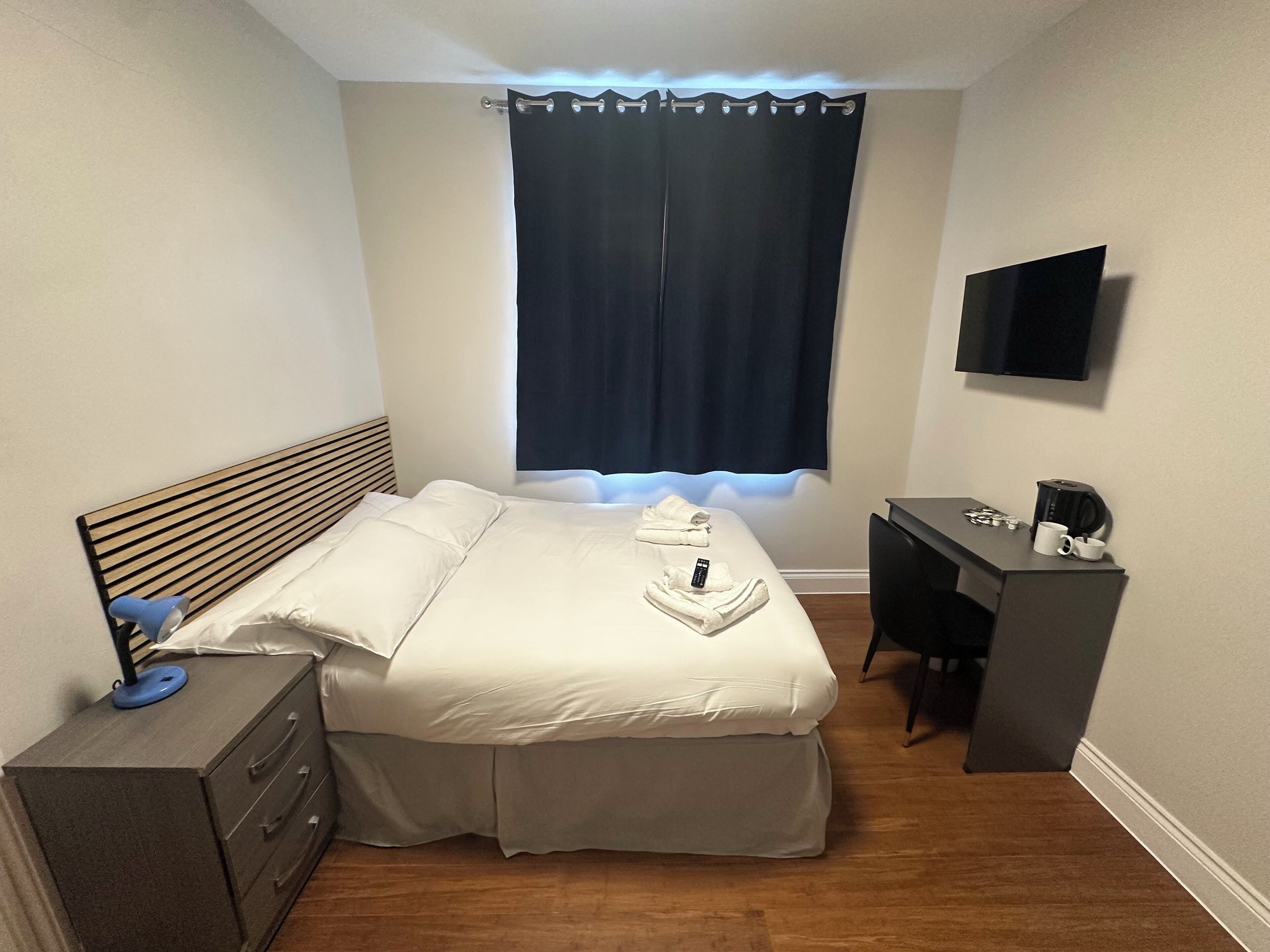 A neatly arranged bedroom with a bed, a desk, a television, and a window with curtains.