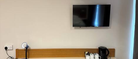 A hotel room with a wooden desk, a black chair, a television mounted on the wall, a coffee maker, and wicker storage drawers.