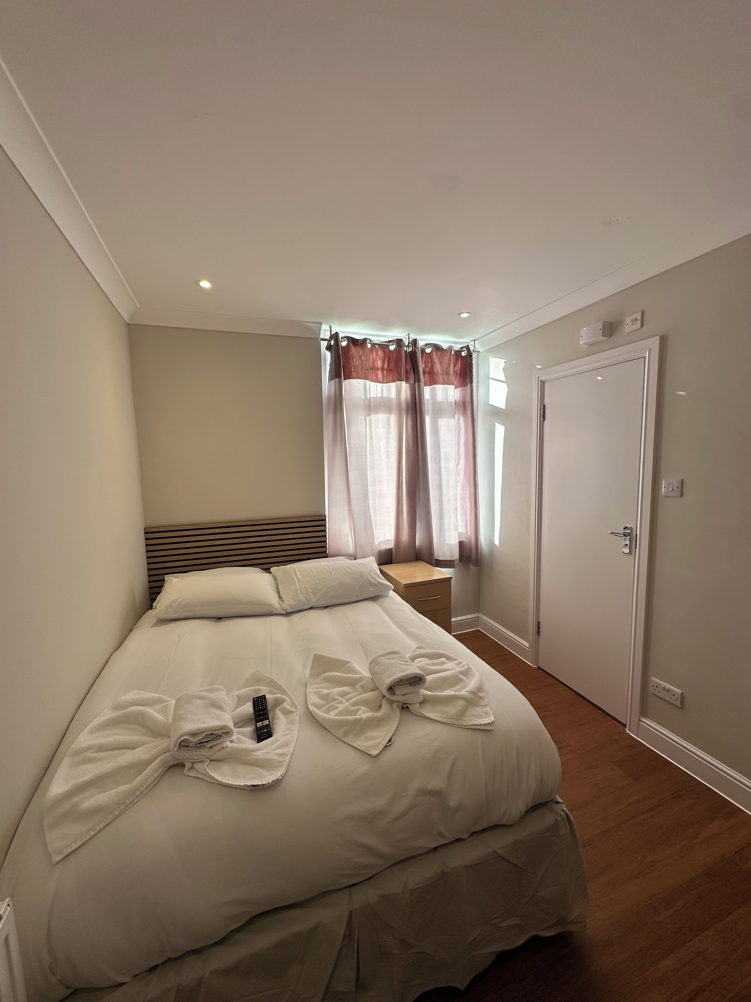 A neatly made bed with towels, a bedside table, a nightstand, a window with curtains, and a door.