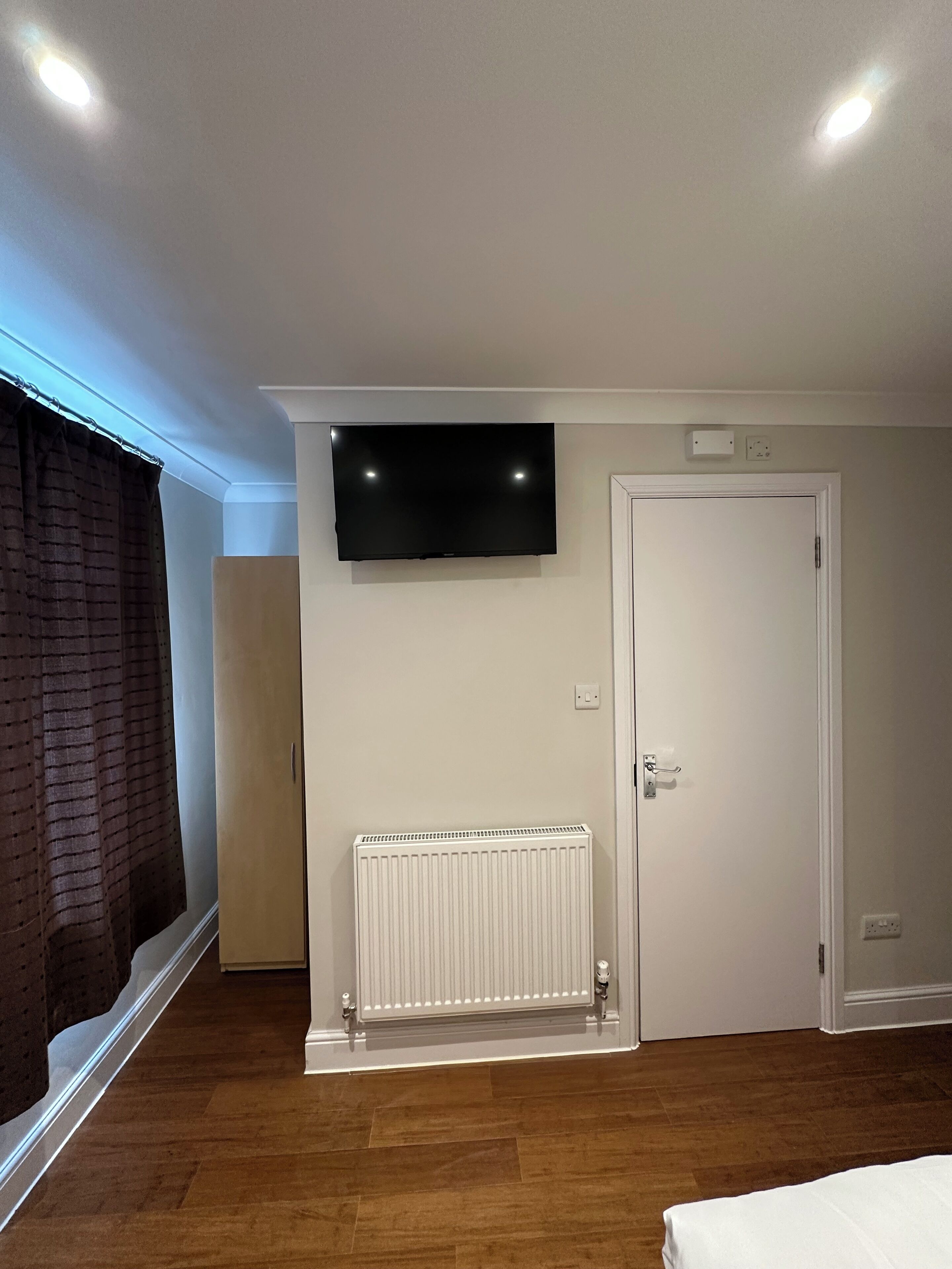 Ein Zimmer mit flachbildfernseher an der Wand, einer weißen Heizung, einer weißen Tür und einem Fenster mit dunklen Vorhängen.
