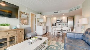 Una sala de estar moderna con un sofá, una mesa de centro y un televisor montado en la pared. Cuenta con una zona de cocina con armarios blancos y una mesa de comedor.