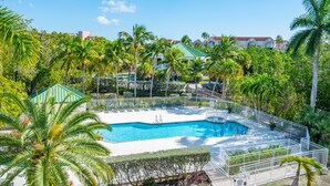 A swimming pool surrounded by palm trees and a white fence, with a clear blue sky in the background. - The Grand Cayman | Beautifully Remodeled Key West! (Key West)