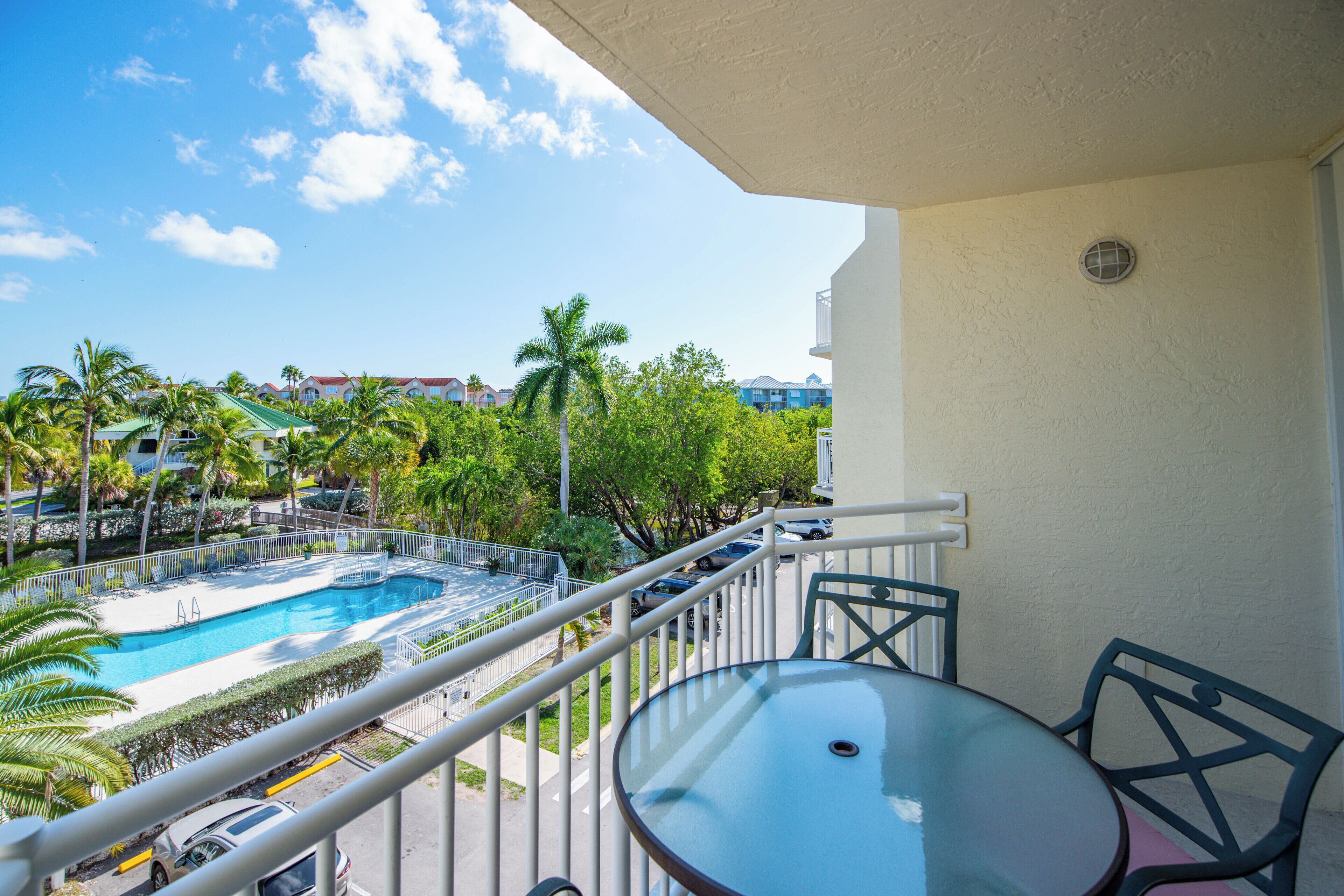 En balkon med et glasbord og stole, der udover et swimmingpool og udsigt over et resortområde med palmer og bygninger.