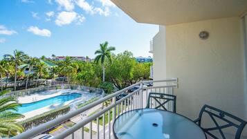 En balkon med et glasbord og stole, der udover et swimmingpool og udsigt over et resortområde med palmer og bygninger.