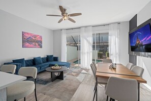 A modern living room with a blue sectional sofa, a dining table with chairs, a ceiling fan, and a TV on the wall. - Dream Stay|Private Pool & Resort Access| 5min to Parks (Kissimmee)