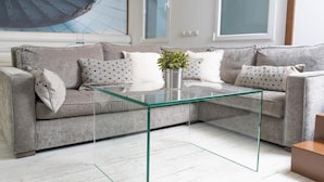 Ein modernes Wohnzimmer mit einem grauen Sofa, gemusterten Kissen, einem Glastisch und einem großen, abstrakten Gemälde an der Wand.