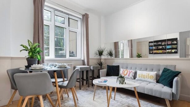 Uma sala de estar moderna com área de jantar, um sofá cinza com almofadas decorativas, uma mesa de centro e uma grande janela com cortinas.