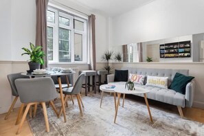 Un salon moderne comprenant un coin repas, un canapé gris agrémenté de coussins décoratifs, une table basse et une grande fenêtre avec des rideaux.