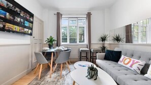 Uma sala de estar moderna com um sofá cinzento, uma mesa de centro branca e um espaço de jantar com uma mesa pequena e cadeiras.