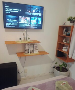 Een woonkamer met een flatscreen televisie aan de muur, waarop een programma wordt vertoond. Daaronder bevinden zich twee houten planken met diverse objecten, waaronder een klein plantje en een decoratieve doos. Op de vloer, naast de tv-standaard, staat een plant in pot.
