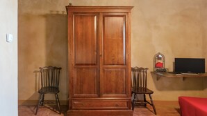A room with a wooden wardrobe, two chairs, a small table with a mirror, and a television.