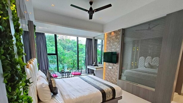 A modern bedroom with a large bed, a ceiling fan, a TV mounted on the wall, and a view of greenery outside. - Melody Boutique Hotel (Thane)