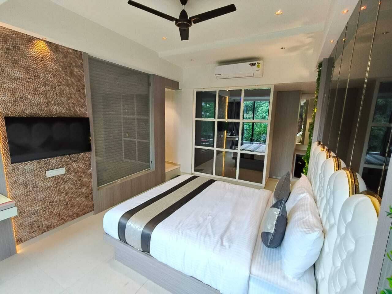 A modern bedroom with a large bed, a ceiling fan, a TV mounted on a textured wall, and a view of greenery through a window.