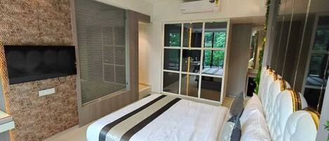 A modern bedroom with a large bed, a ceiling fan, a TV mounted on a textured wall, and a view of greenery through a window.