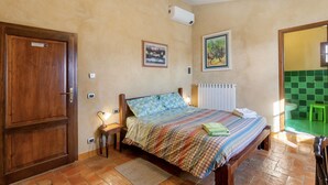 Un dormitorio con puerta de madera, una cama con colcha estampada, aire acondicionado instalado en la pared y un baño con azulejos verdes visible a través de un umbral abierto.