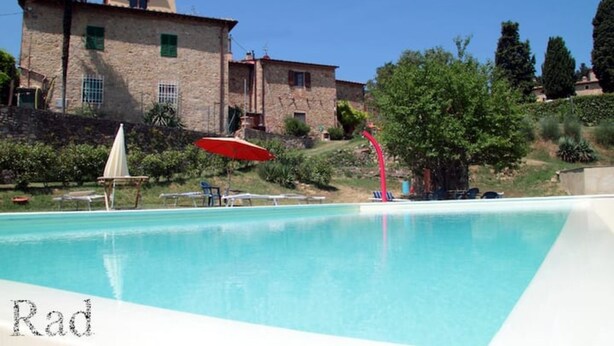 Una piscina con sillones de descanso y sombrillas, un edificio de piedra con contraventanas verdes y un cielo azul despejado.