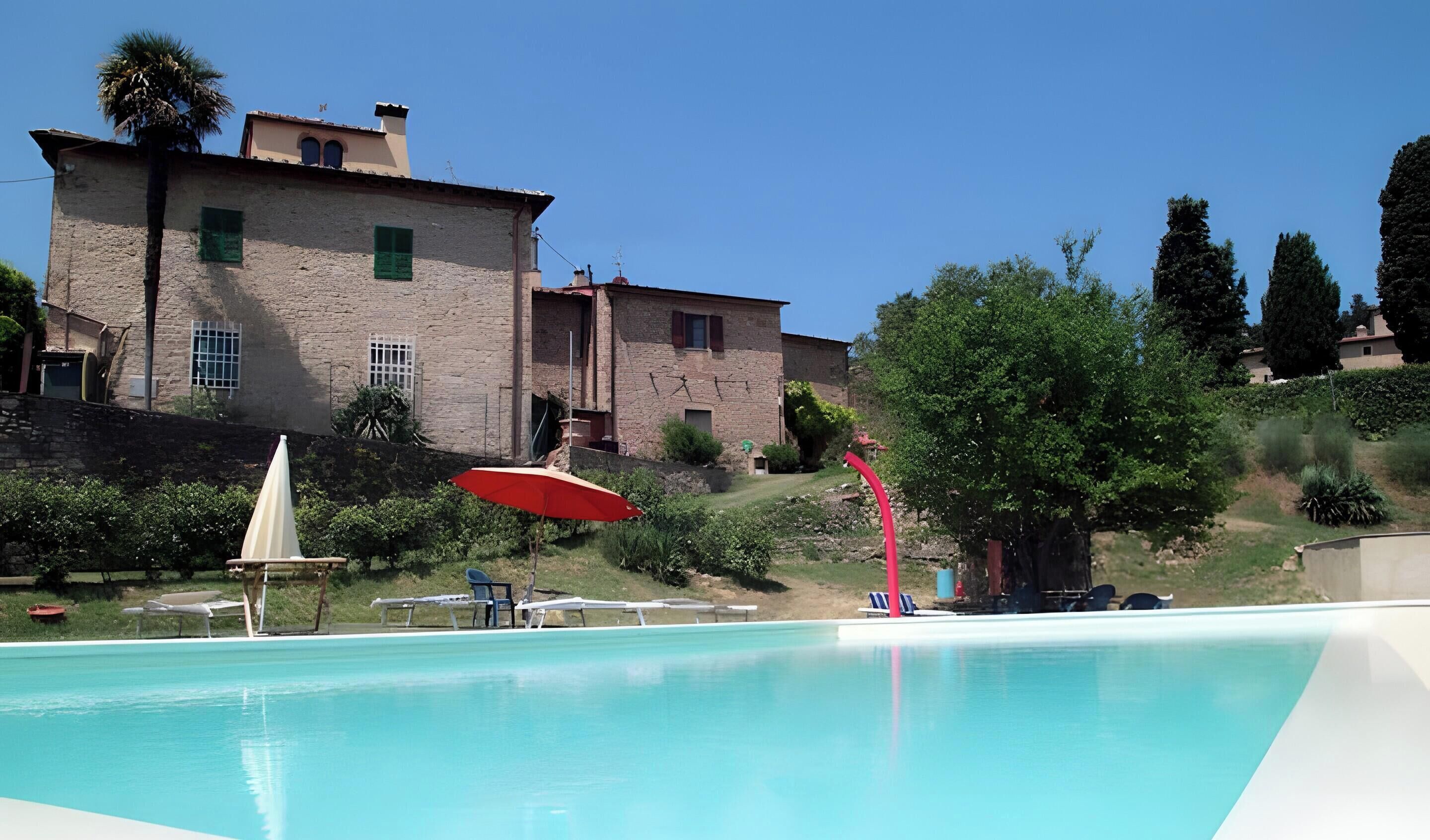 Une piscine avec des chaises longues et un parasol rouge, entourée de verdure et d’un bâtiment en pierre.