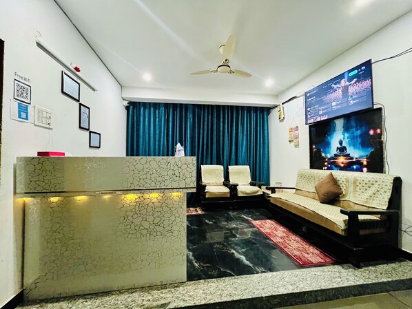 A living room with a sofa set, a flat-screen TV, a ceiling fan, and a blue curtain. - AVR MOTEL (Varanasi)