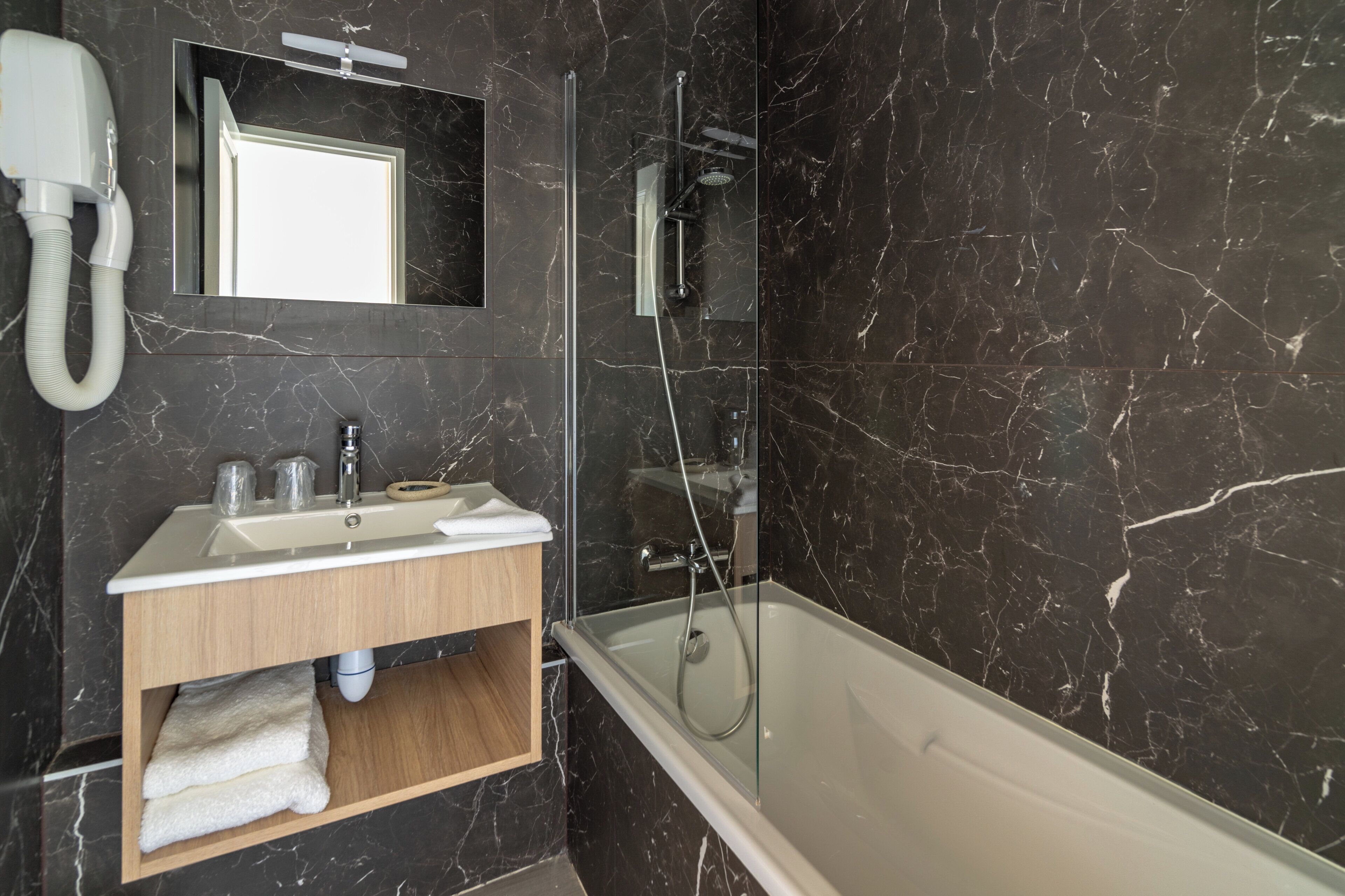 Un baño moderno con pared de mármol, un lavabo blanco, un mueble de tocador de madera, ducha con cerramiento de vidrio y una bañera blanca.