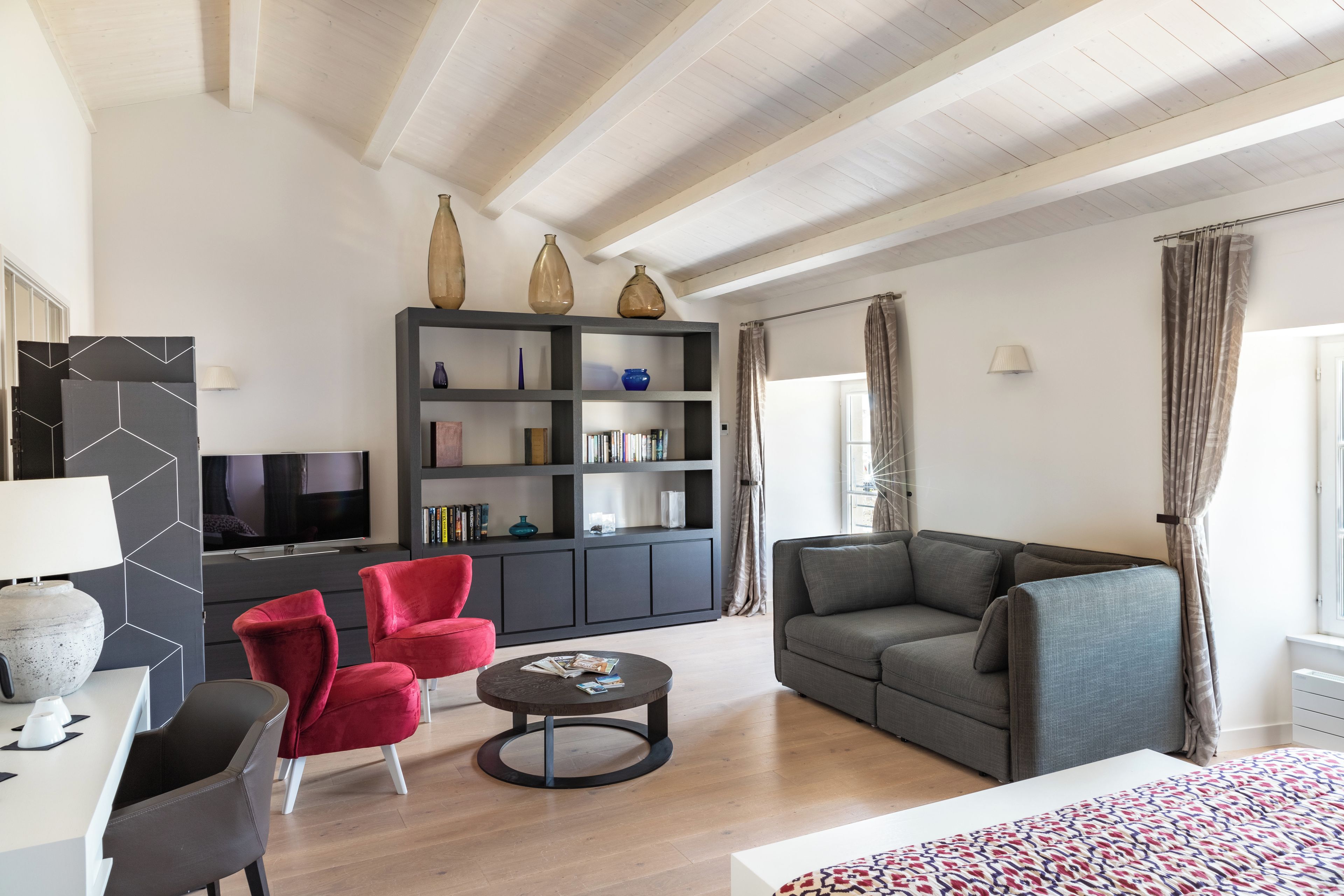 Ein modernes Wohnzimmer mit einem grauen Sofa, roten Sesseln, einem Couchtisch, einem Fernseher und einem Bücherregal mit dekorativen Vasen.