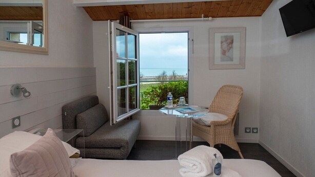 Ein Schlafzimmer mit einem Bett, einem grauen Sofa, einem Korbstuhl, einem kleinen Tisch, einem Fernseher und einem Fenster mit Meerblick.