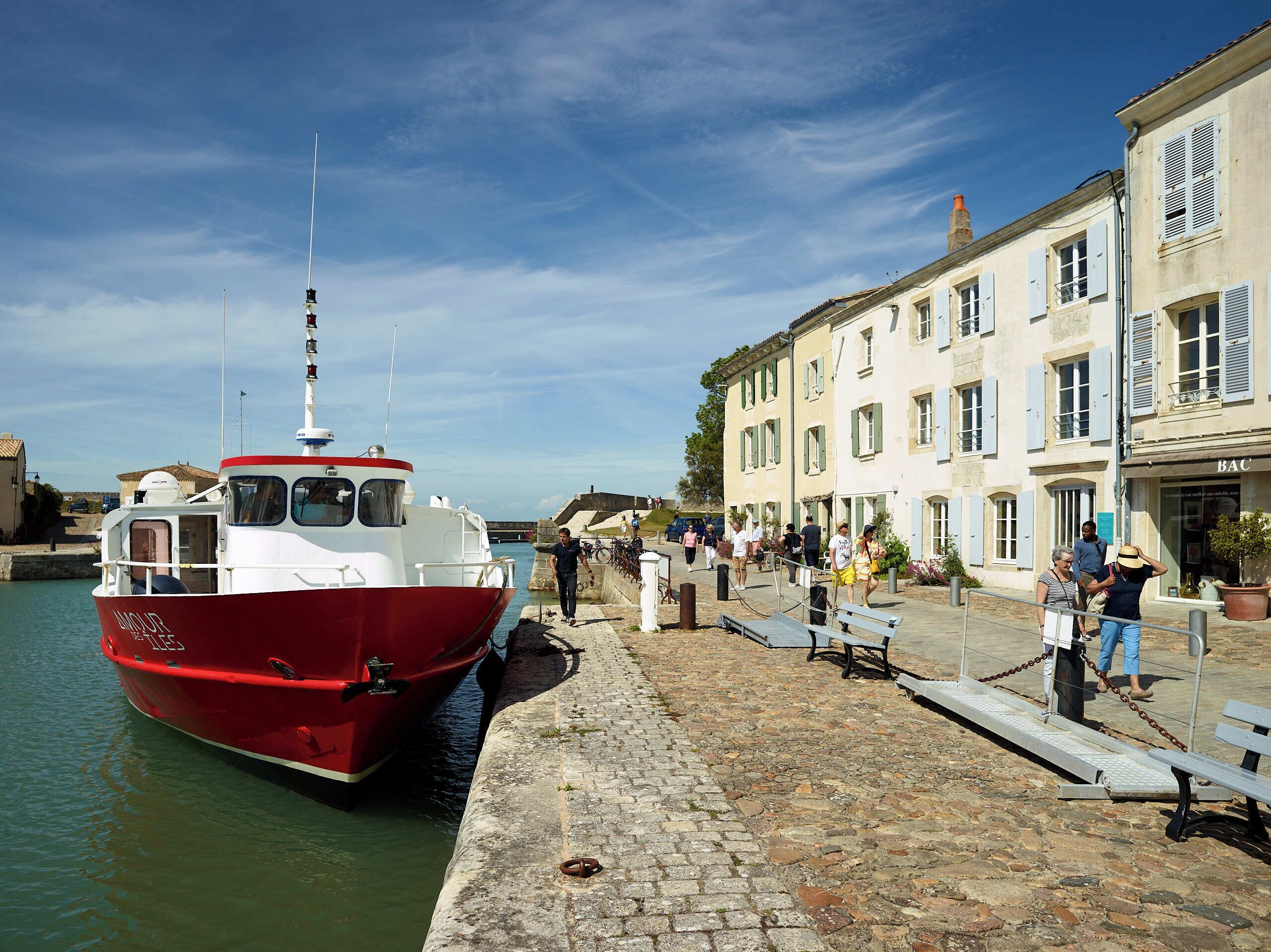 A red boat docked at a waterfront with cobblestone walkways and buildings with shutters.