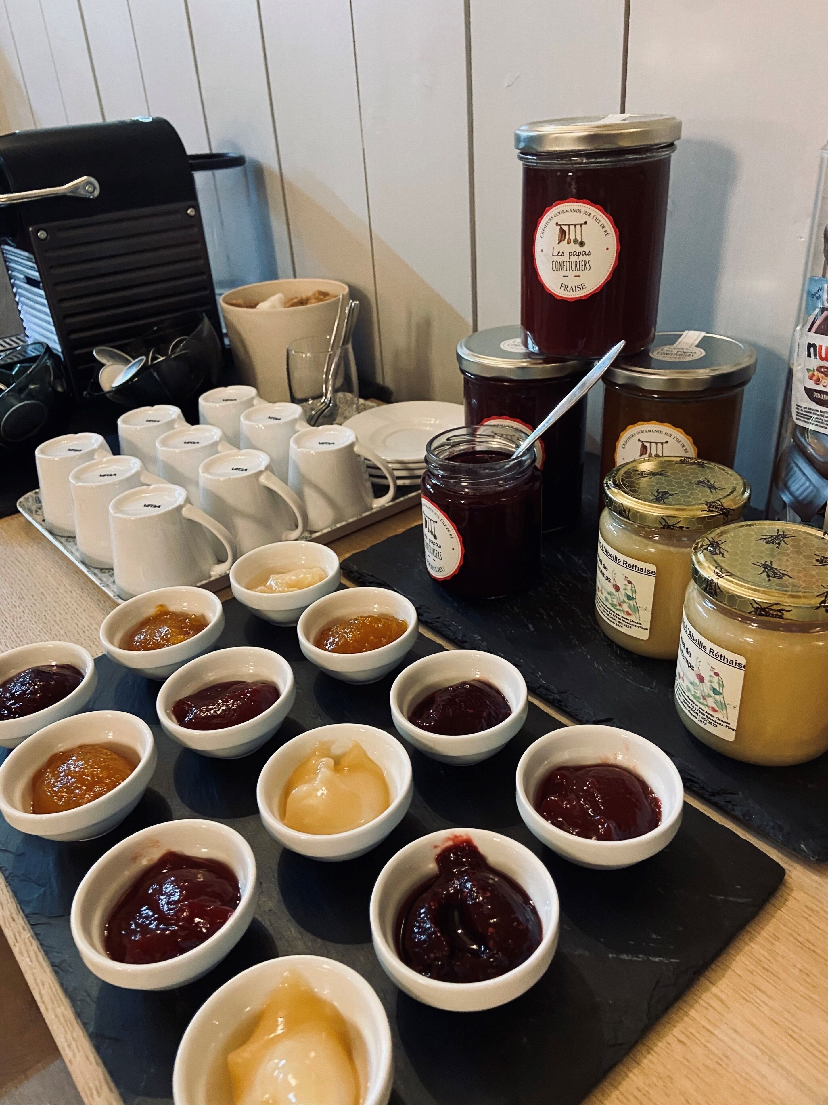A variety of condiments and jars on a tray, with a coffee machine and cups in the background.