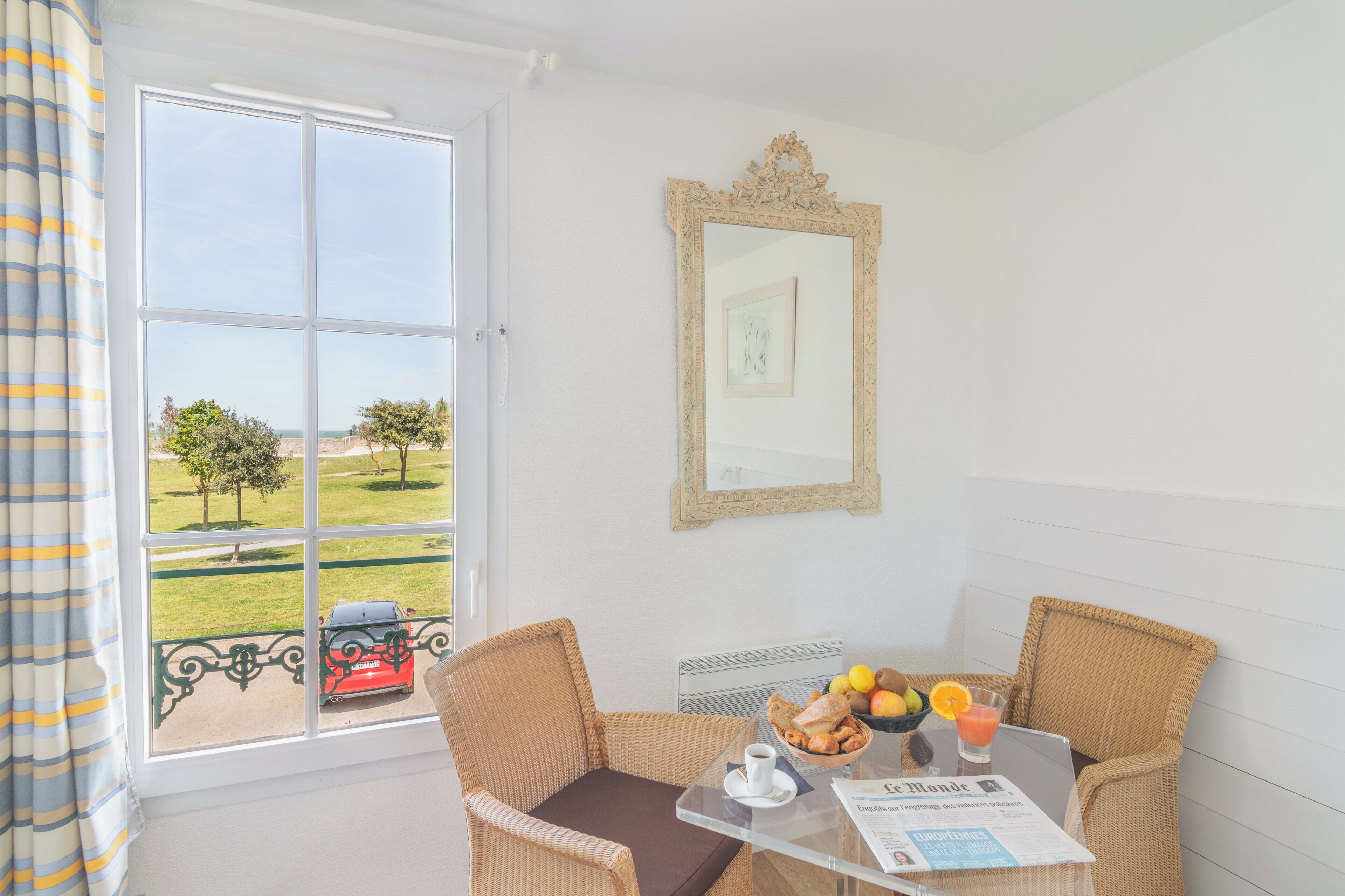 Una habitación con vista a un paisaje verde y un coche rojo, sillas de mimbre, una mesa de vidrio con alimentos para el desayuno y un cuadro en la pared.