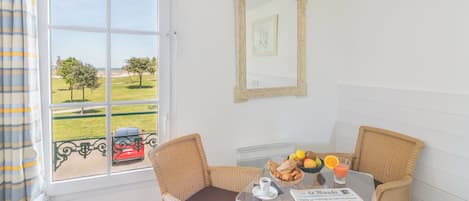 A room with a window view of a green landscape and a red car, wicker chairs, a glass table with breakfast items, and a framed picture on the wall.