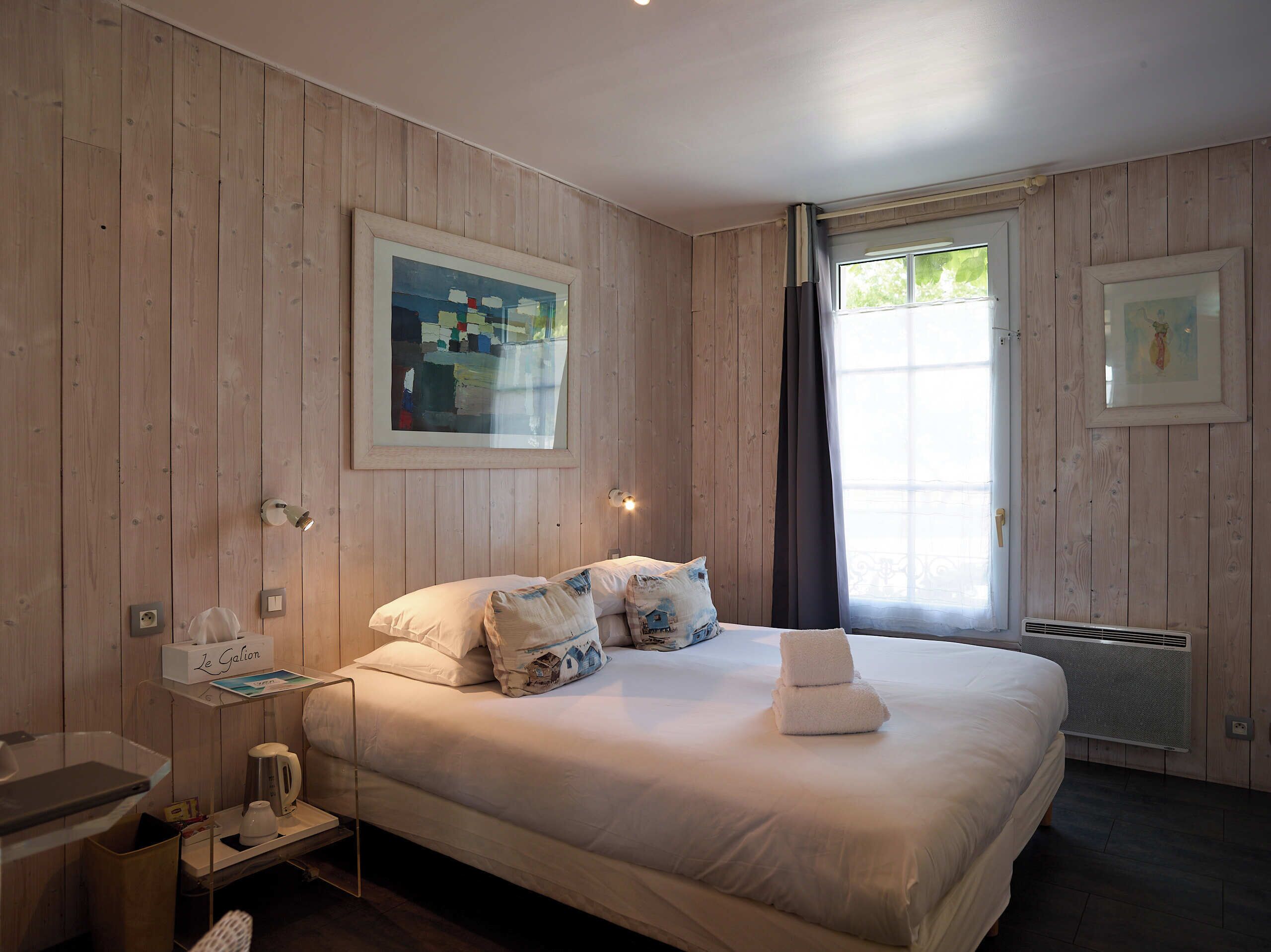 A hotel room with wooden panel walls, a bed with white linens, a painting, a window with curtains, and a bedside table with a lamp.