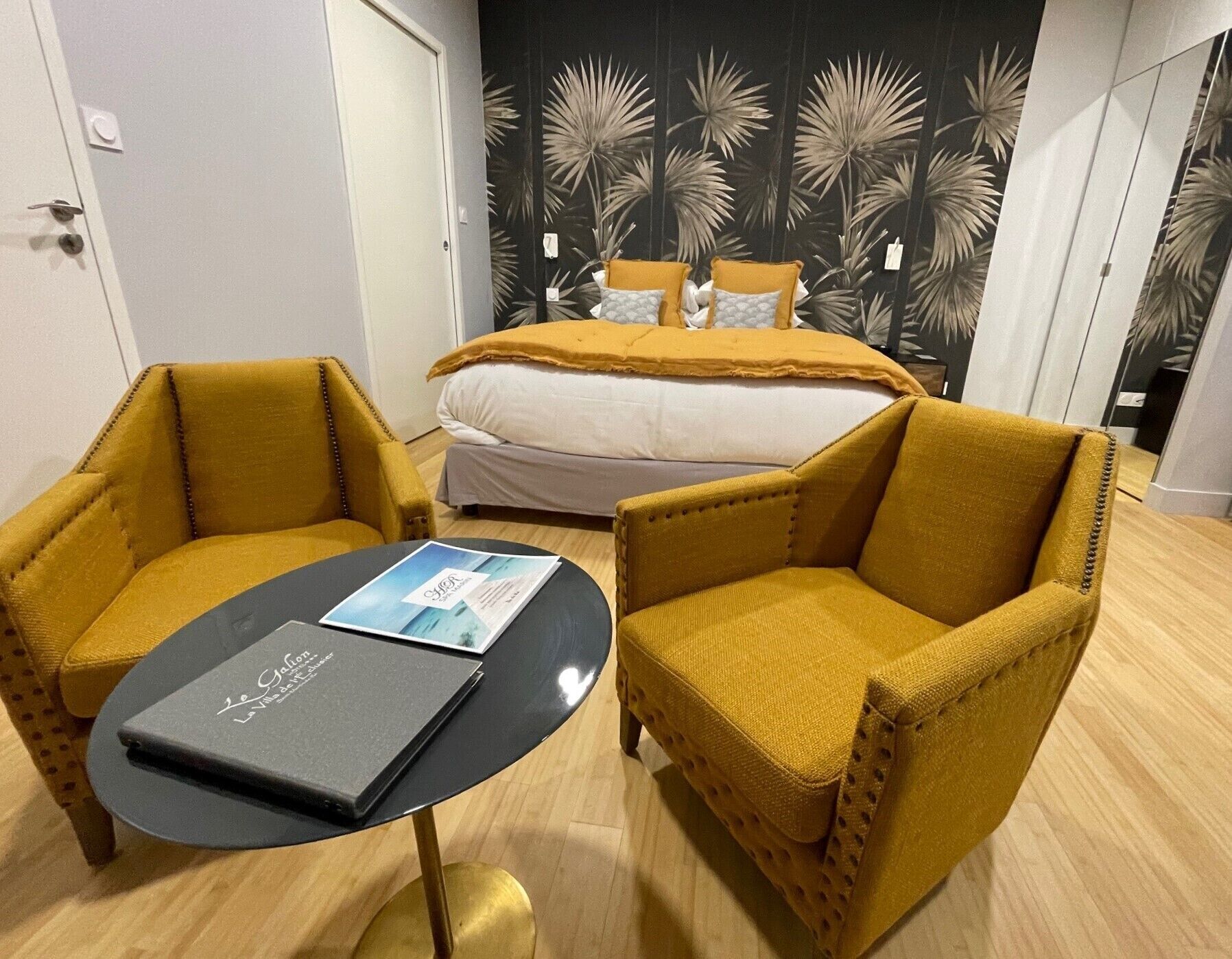 Habitación de hotel con cama, dos sillones amarillos, una mesita con una revista y un papel tapiz tropical.