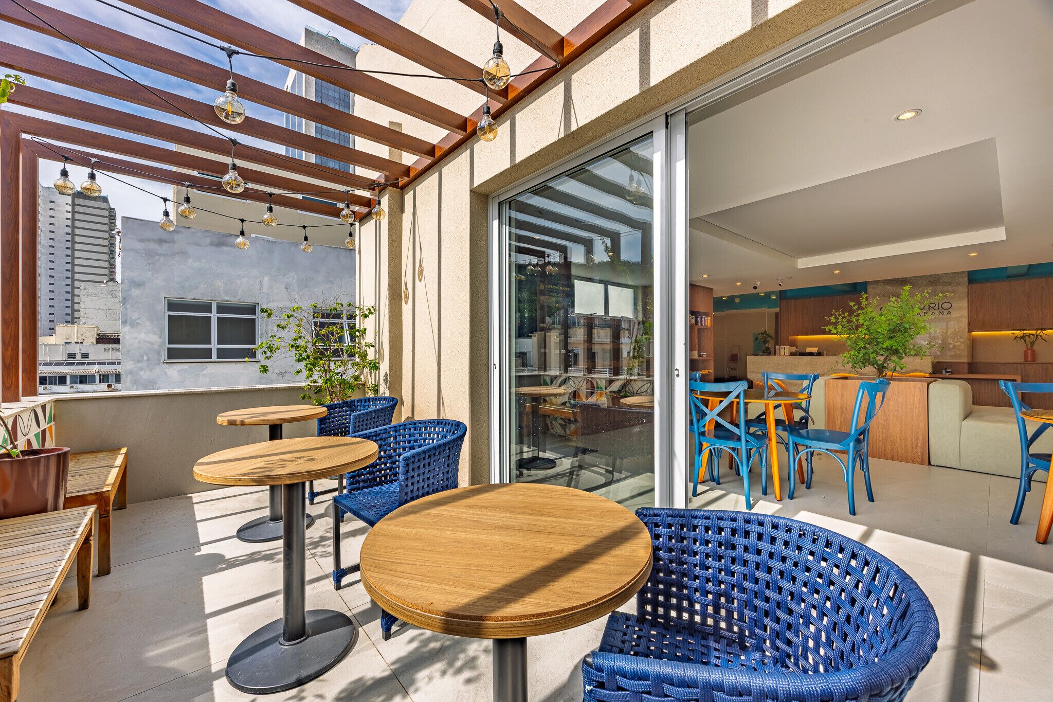 An outdoor seating area with wooden tables and blue wicker chairs, a wooden pergola with hanging lights, and a glass door leading to an indoor space.