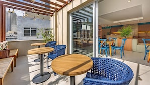 Un'area esterna con tavoli di legno e sedie in vimini blu, un pergolato in legno con luci a sospensione e una porta vetrata che dà su un ambiente interno.