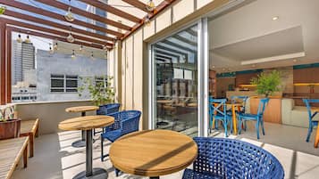 An outdoor seating area with wooden tables and blue wicker chairs, a wooden pergola with hanging lights, and a glass door leading to an indoor space.