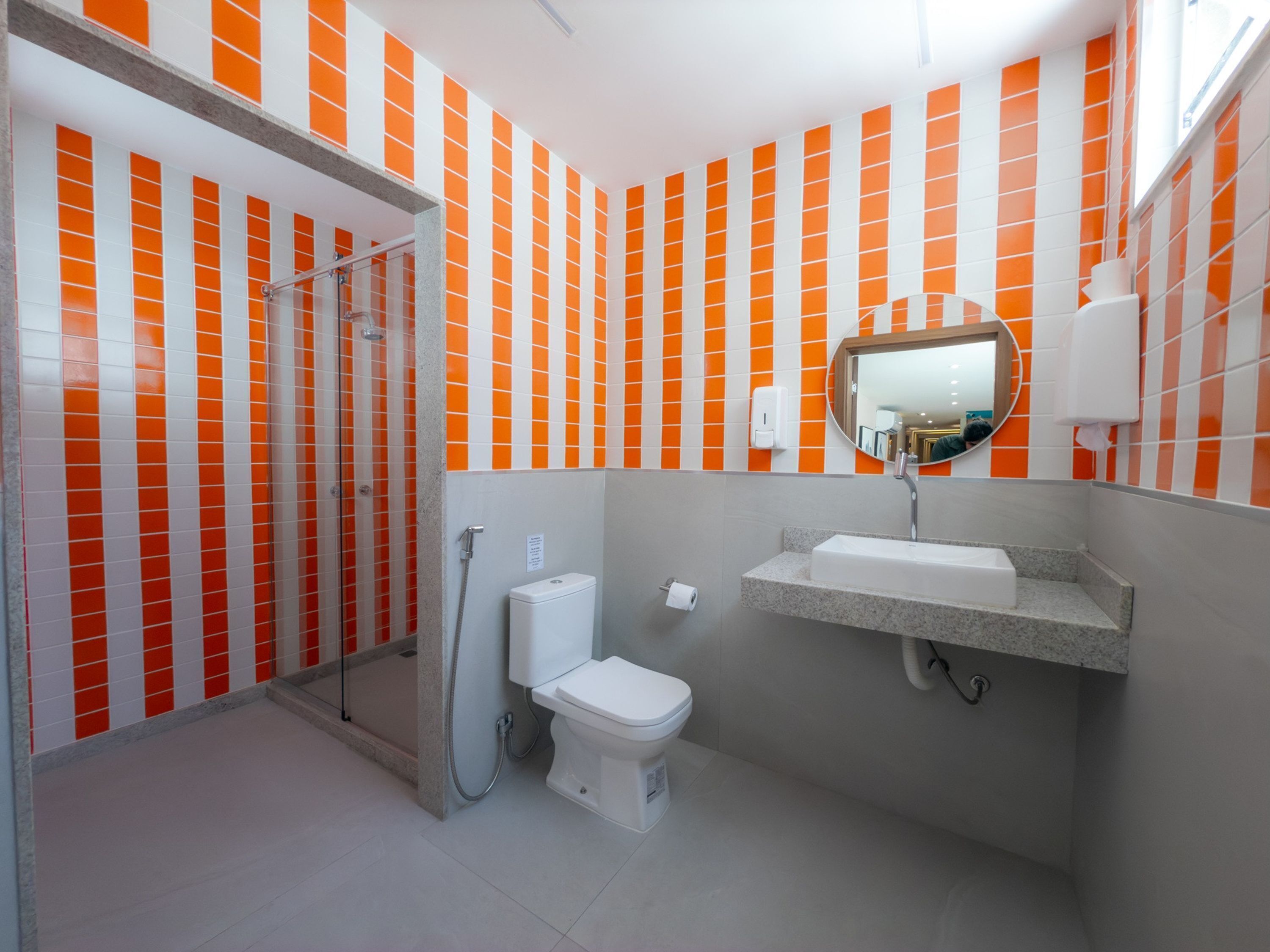 Un baño con paredes de azulejos naranja y blanco, cabina de ducha de vidrio, inodoro y lavamanos con espejo.