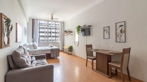 Una sala de estar moderna con un sofá, una mesa de comedor con sillas, un televisor montado en la pared y un ventanal con cortinas.