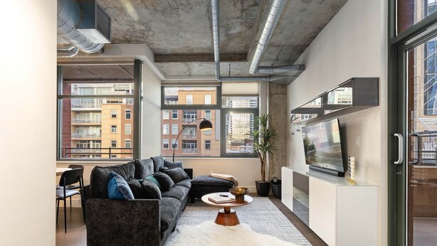 A modern living room with a grey sofa, a white rug, a TV, and large windows offering a view of buildings outside. - Union Station downtown and gameday retreat! (Denver)