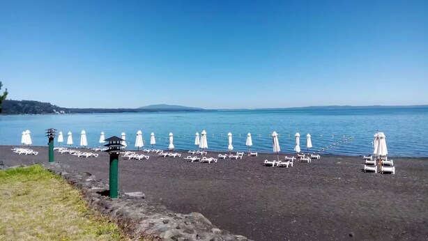 Une plage bordée de rang ées de chaises longues et de parasols blancs, un rivage rocheux et une vue dégagée sur l’eau et les collines lointaines.