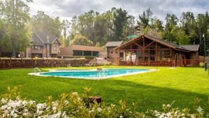 Une piscine entourée d’une pelouse bien entretenue, avec un chalet en bois et un bâtiment plus grand en arrière-plan.