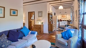 Una sala de estar con sofá, sillón y una mesa de centro. Se observa una zona de cocina al fondo con armarios y una mesa de comedor.