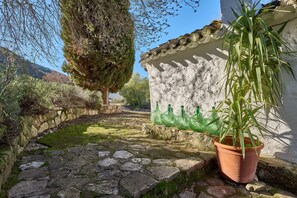 Un chemin de pierres mène à un bâtiment blanc doté d’un toit en tuiles, entouré de verdure et surplombé d’un ciel dégagé.