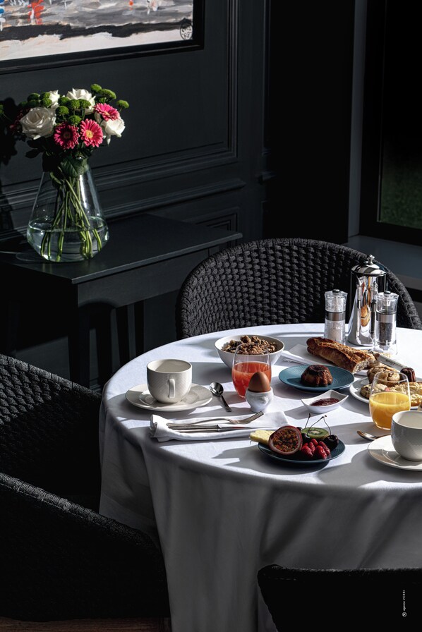 Una mesa de desayuno con diversos alimentos, un jarrón con flores y vista al exterior.