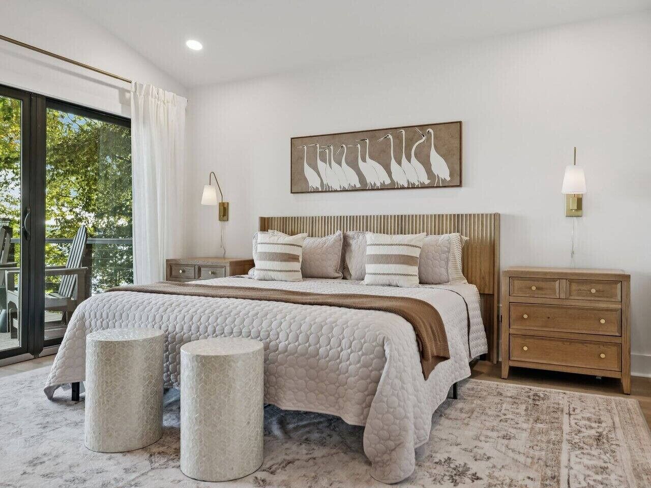 A bedroom with a large bed, two bedside tables, a view through a glass door, and a painting on the wall.