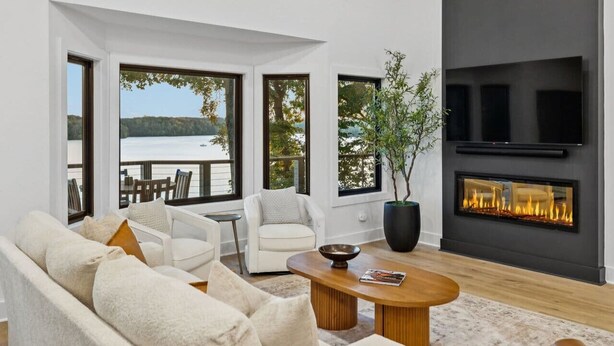 Una sala de estar moderna con chimenea, sofás blancos, una mesa de centro y amplios ventanales con vistas a un lago.