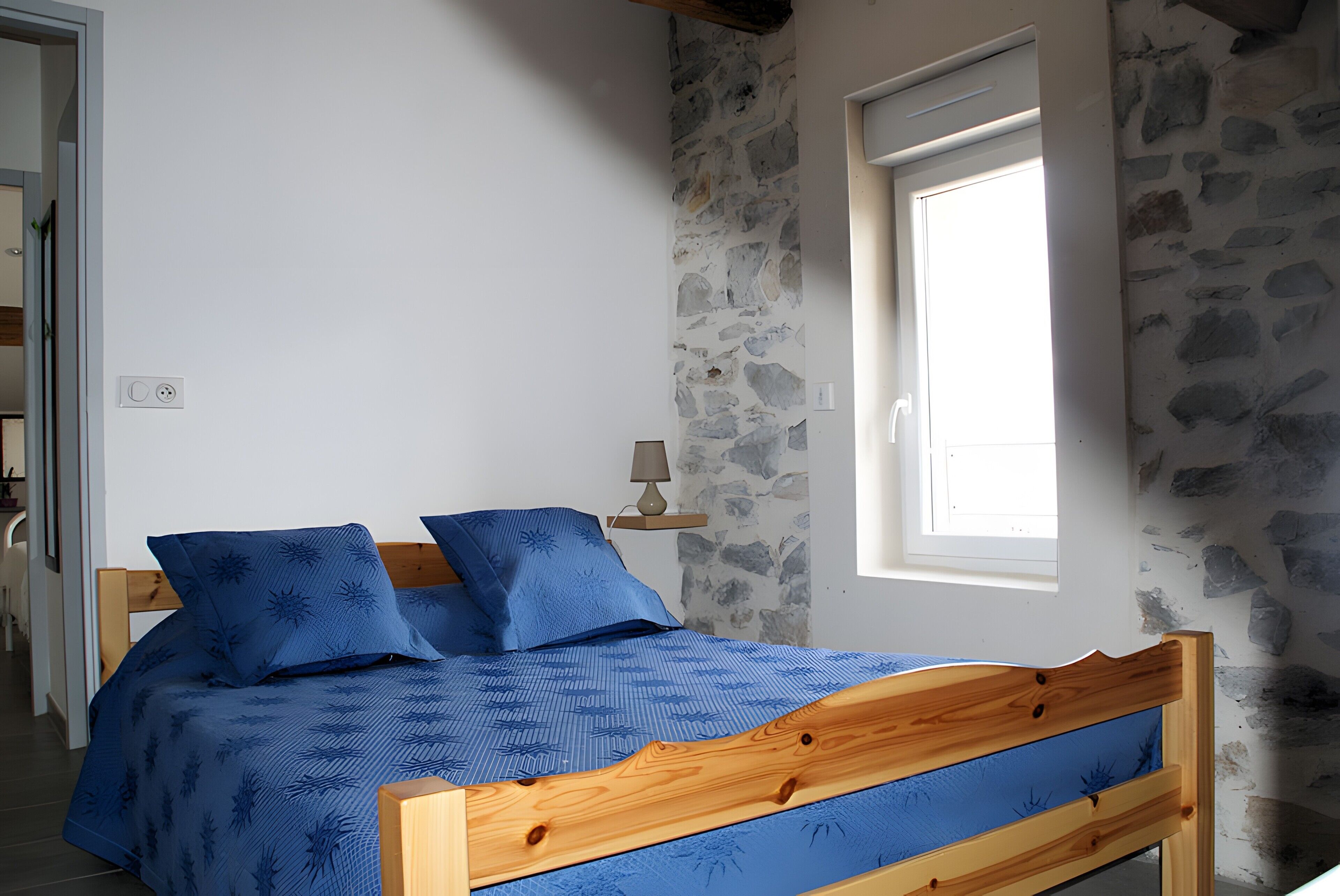 Un lit avec un cadre en bois et une literie bleue, un mur en pierre, une fenêtre et une petite table de chevet avec une lampe.