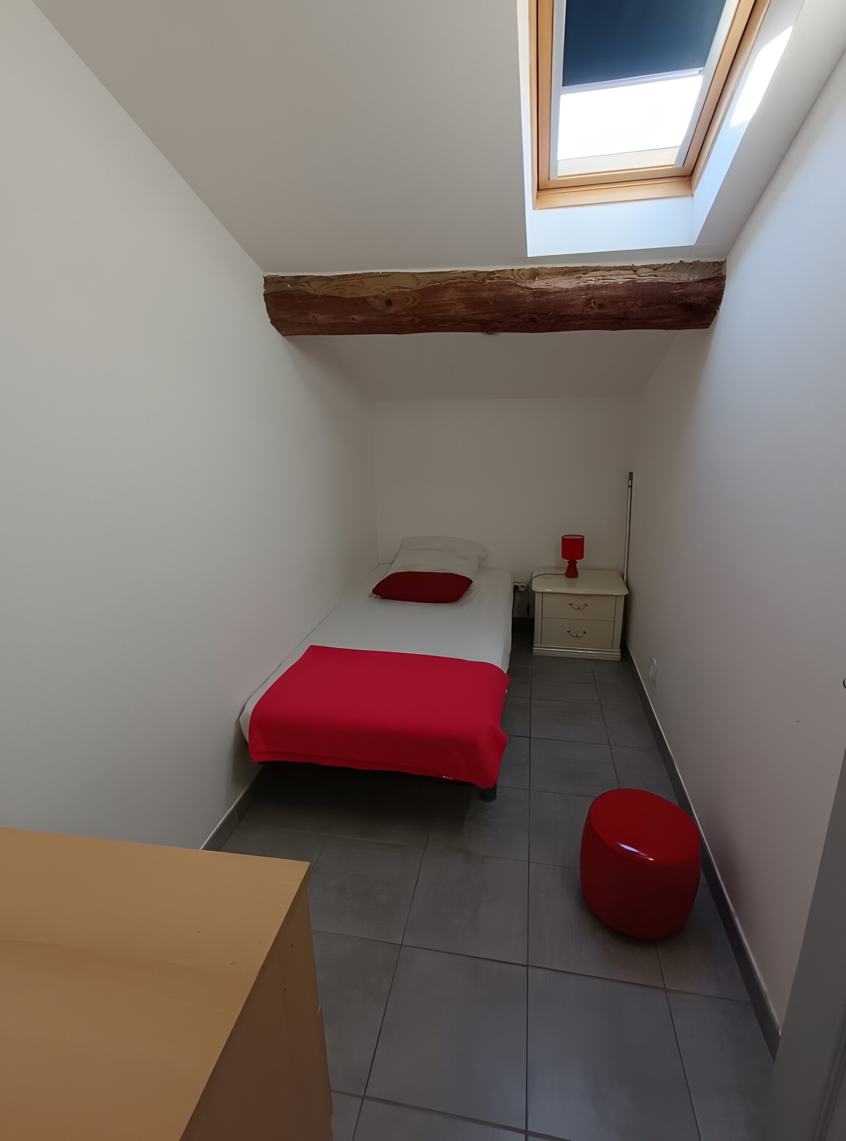 A small, narrow room with a skylight, a bed with red bedding, a bedside table with a lamp, and a red stool.