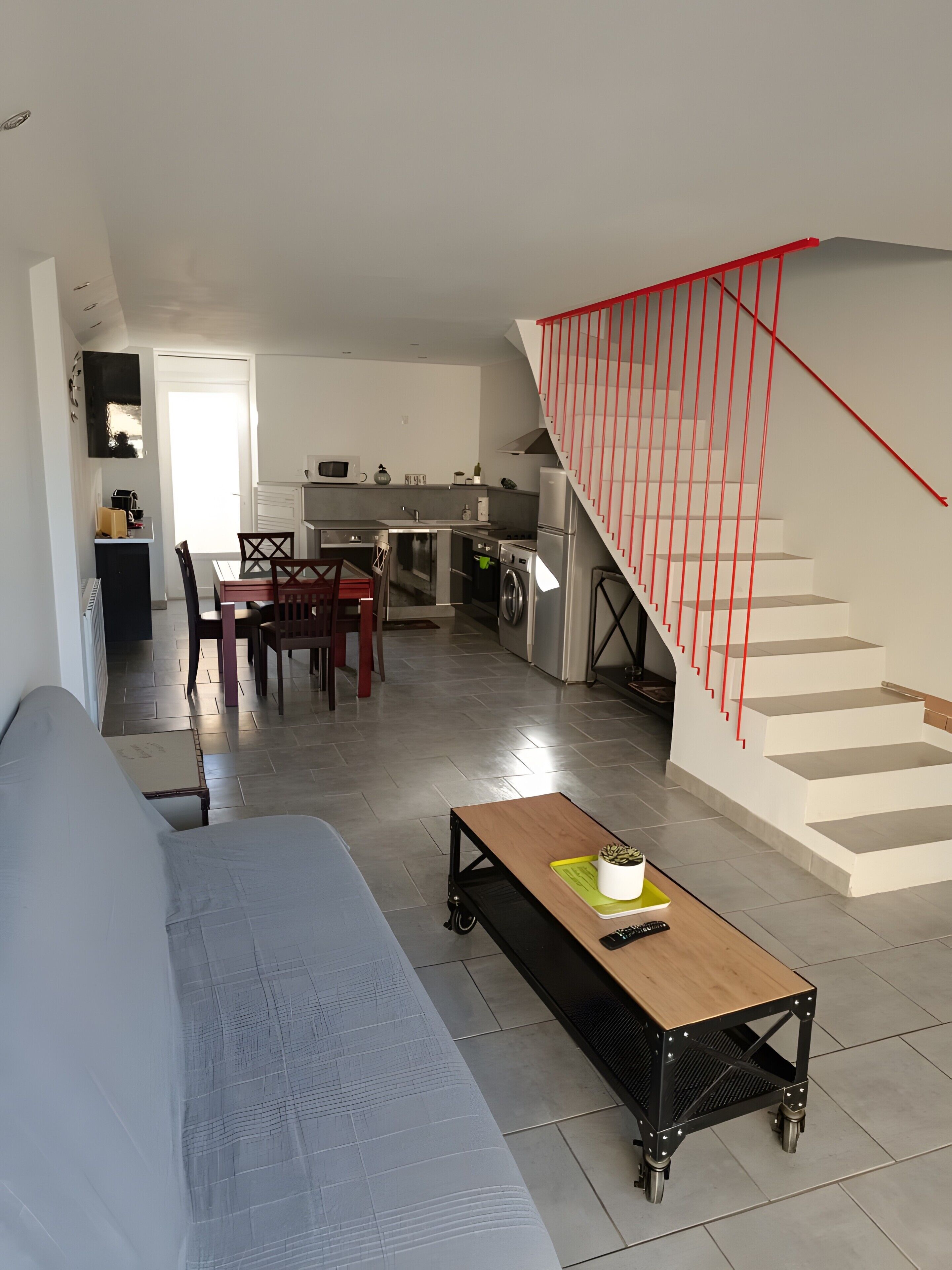 Ein Wohnzimmer mit Sofa, Couchtisch, Essbereich mit Stühlen, Küche inklusive Geräte und einer Treppe mit roten Geländern.