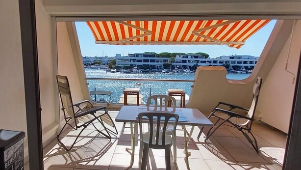 A balcony with a table and chairs, a striped umbrella, and a view of a marina with boats and buildings. - “Sea & Sun” Apartment with Private Terrace (Port Camargue)