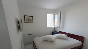 A bedroom with a bed, a bedside table, a lamp, a picture on the wall, and a window. - “Sea & Sun” Apartment with Private Terrace (Port Camargue)